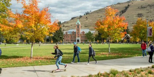 A picture of the University of Montana campus quad in Fall. Students walk and skateboard down a path surrounded by Fall foliage. 